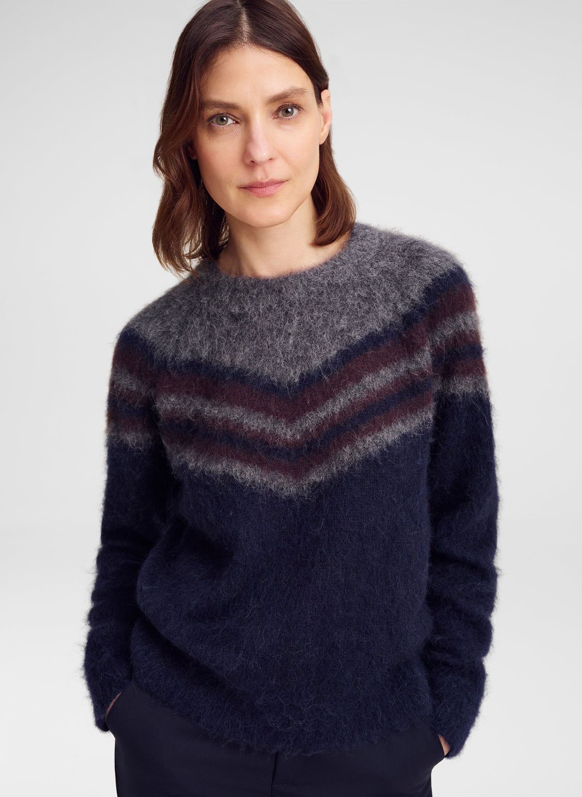 A woman wearing a fuzzy knit crewneck sweater with a chevron pattern in navy blue, dark gray, and burgundy, photographed against a light gray studio background.