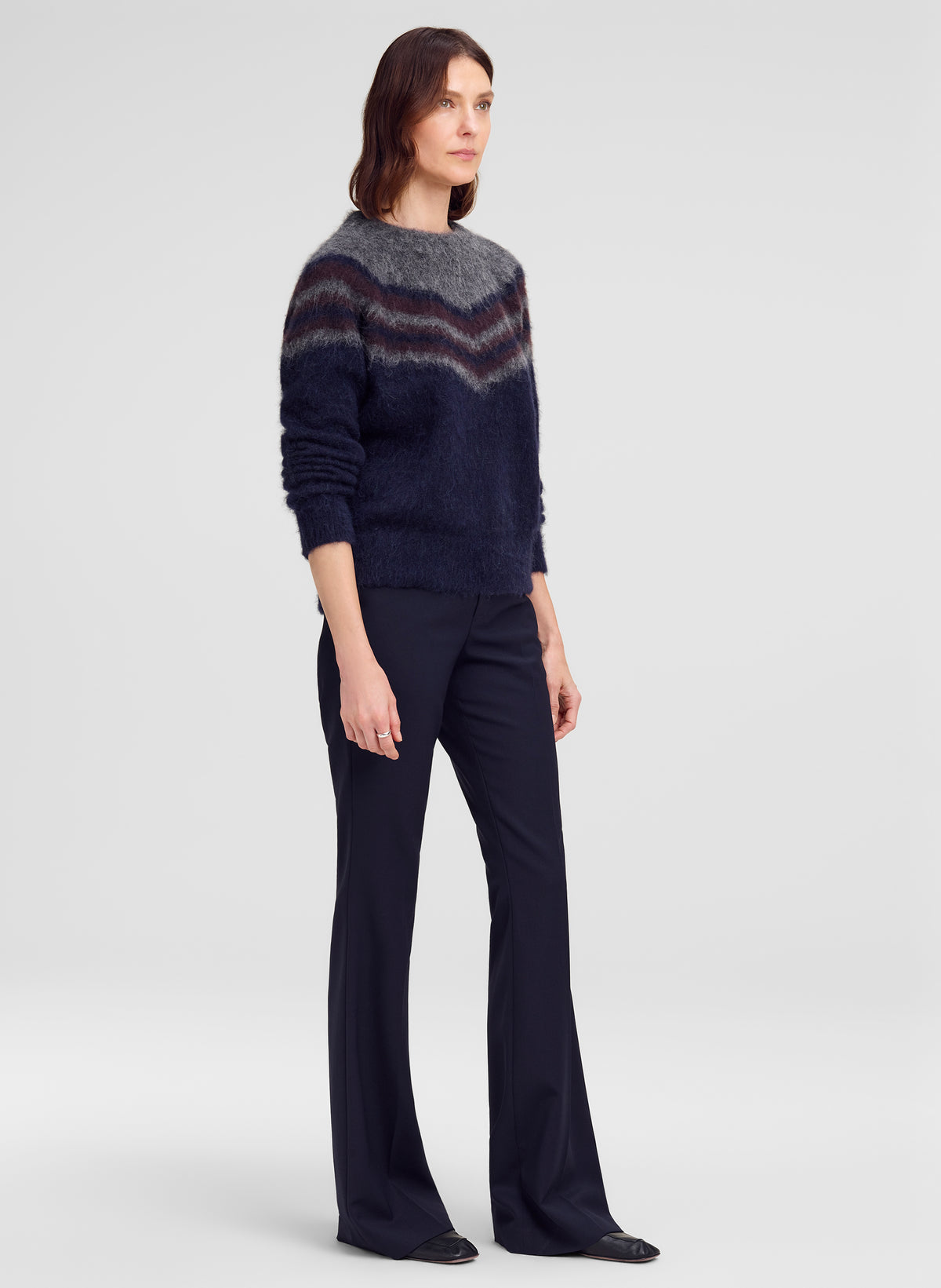 A woman modeling a dark navy knit sweater with a gray and burgundy chevron pattern across the chest, paired with dark navy trousers, against a light gray studio background.
