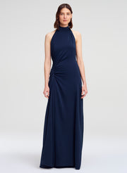 A woman modeling a sleeveless navy blue halter-neck maxi dress with a gathered, draped design on the bodice and skirt, set against a plain light gray studio background.