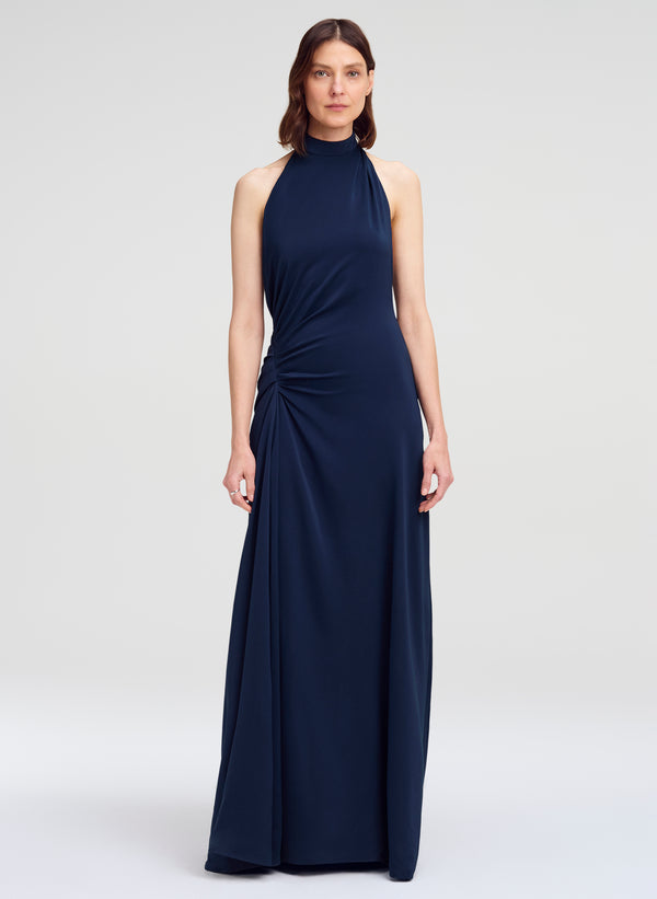 A woman modeling a sleeveless navy blue halter-neck maxi dress with a gathered, draped design on the bodice and skirt, set against a plain light gray studio background.