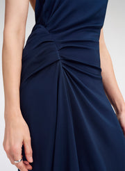 Close-up of a navy blue sleeveless dress with gathered ruching at the waist, worn by a model whose hands and arms are visible against a light gray studio background.