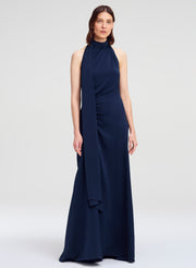 A woman modeling a sleeveless navy blue halter-neck maxi dress with a draped side detail, photographed against a light gray studio background.