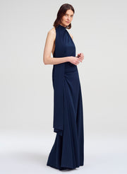 A woman modeling a sleeveless navy blue halter-neck maxi dress with a draped, gathered skirt against a light gray studio background.