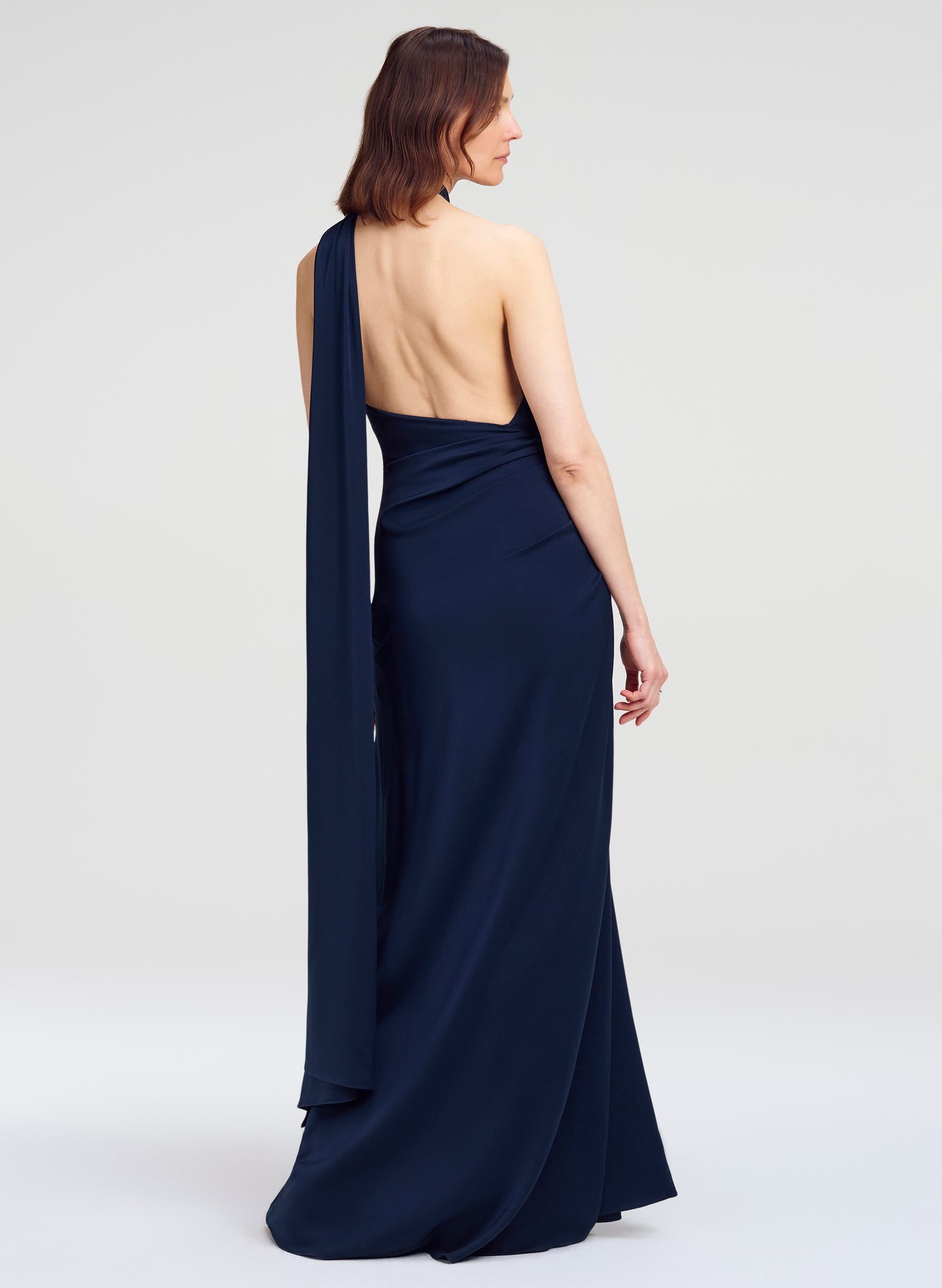 A woman modeling a navy blue one-shoulder floor-length gown from the back, showing a draped open back and long flowing fabric panel, against a light gray studio background.