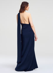 A woman modeling a navy blue one-shoulder floor-length gown from the back, showing a draped open back and long flowing fabric panel, against a light gray studio background.