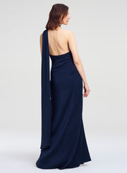 A woman modeling a navy blue one-shoulder evening gown from the back, featuring a draped, open back and a long flowing fabric panel, set against a plain light gray studio background.