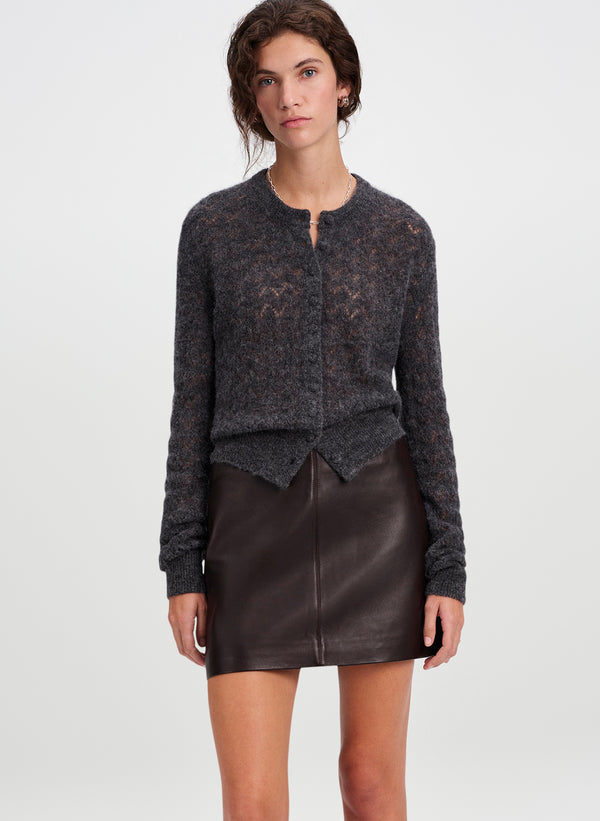 A woman modeling a dark charcoal gray textured knit cardigan with a split neckline, paired with a dark brown leather mini skirt, against a plain light gray studio background.