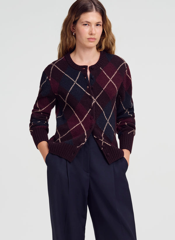 A woman modeling a dark burgundy and navy argyle cardigan sweater with beige diamond lines, paired with navy trousers, against a light gray studio background.