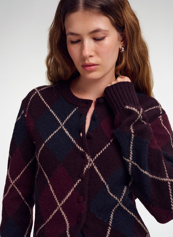 A woman wearing a dark argyle cardigan sweater with burgundy, navy, and beige diamond patterns, photographed against a light gray studio background.