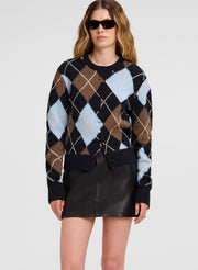A woman modeling a cropped argyle sweater with navy, brown, tan, and light blue diamonds, paired with a black mini skirt against a light gray studio background.