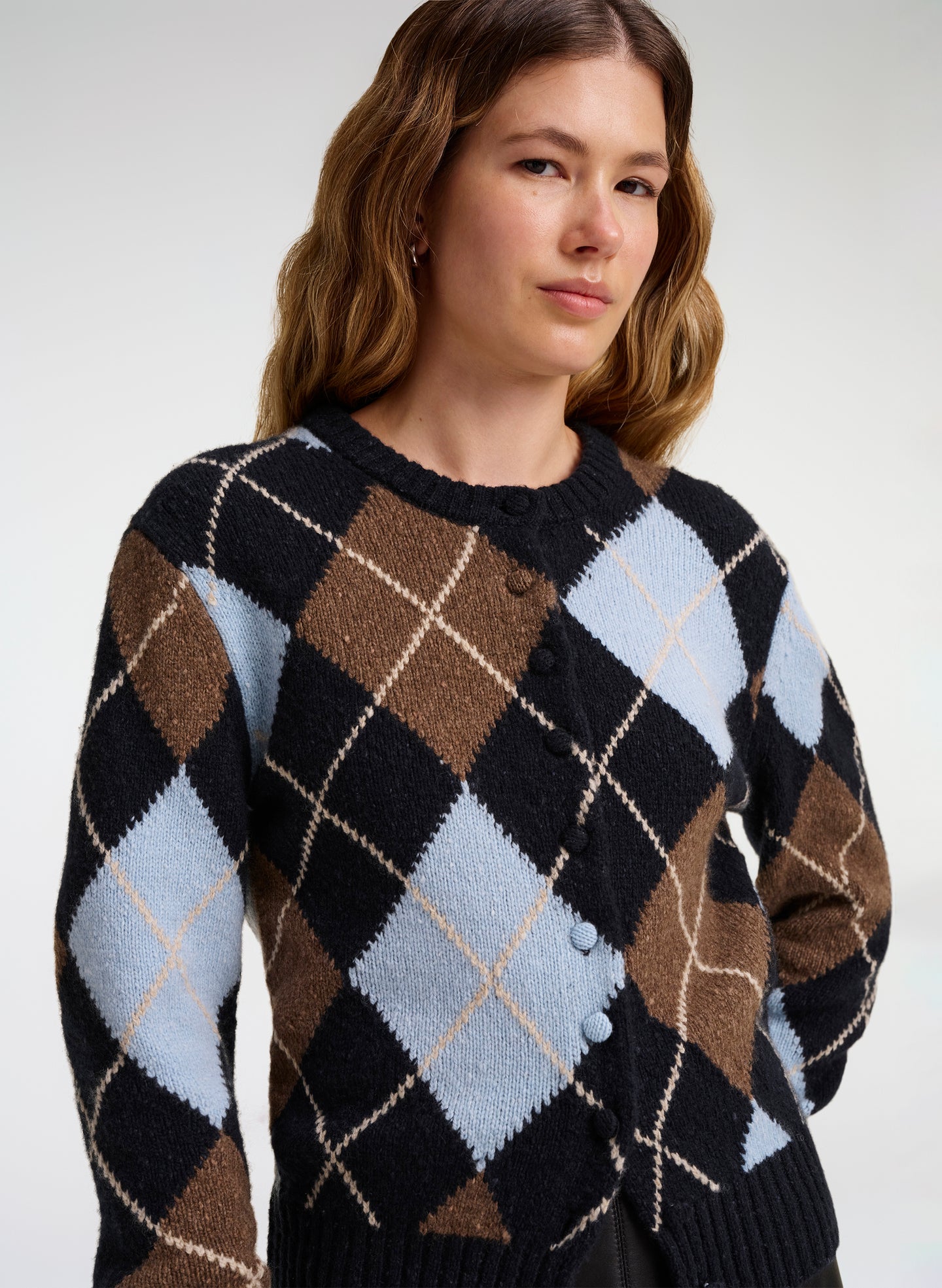 A woman wearing a button-front cardigan with an argyle pattern in black, brown, light blue, and cream, photographed against a plain light gray studio background.