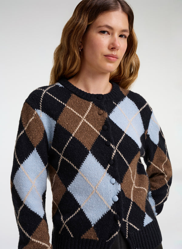 A woman wearing a button-front cardigan with an argyle pattern in black, brown, light blue, and cream, photographed against a plain light gray studio background.