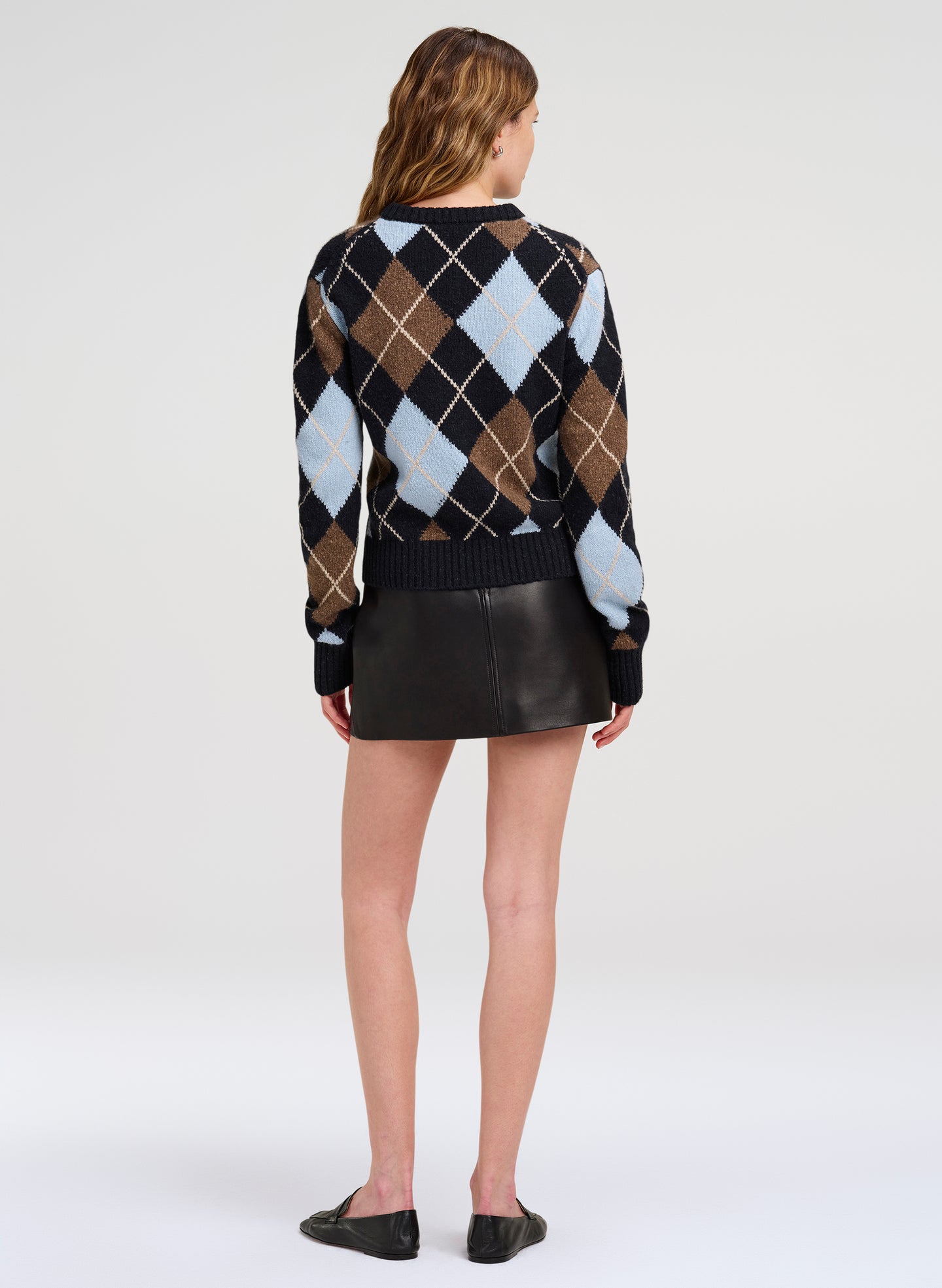A woman shown from the back wearing a black, brown, and light blue argyle sweater with a black mini skirt and black flats, photographed against a light gray studio background.