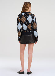 A woman shown from the back wearing a black, brown, and light blue argyle sweater with a black mini skirt and black flats, photographed against a light gray studio background.