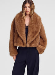 A model wearing a cropped, shaggy brown faux fur jacket over a black top and dark wide-leg trousers, photographed against a light gray studio background.