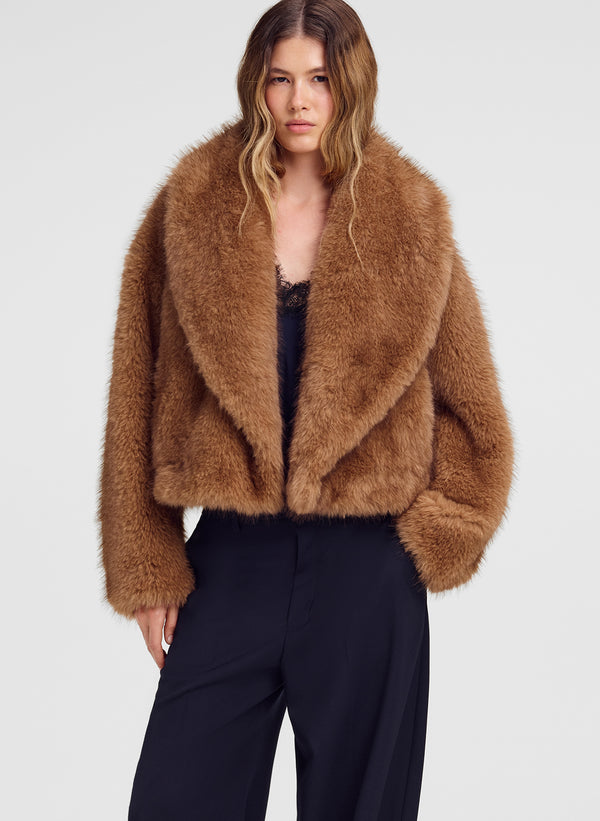 A model wearing a cropped, shaggy brown faux fur jacket over a black top and dark wide-leg trousers, photographed against a light gray studio background.