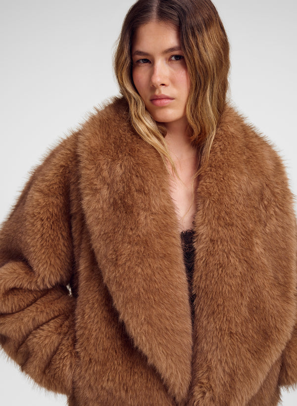 A woman wearing a voluminous, oversized brown faux fur coat against a plain light gray studio background.