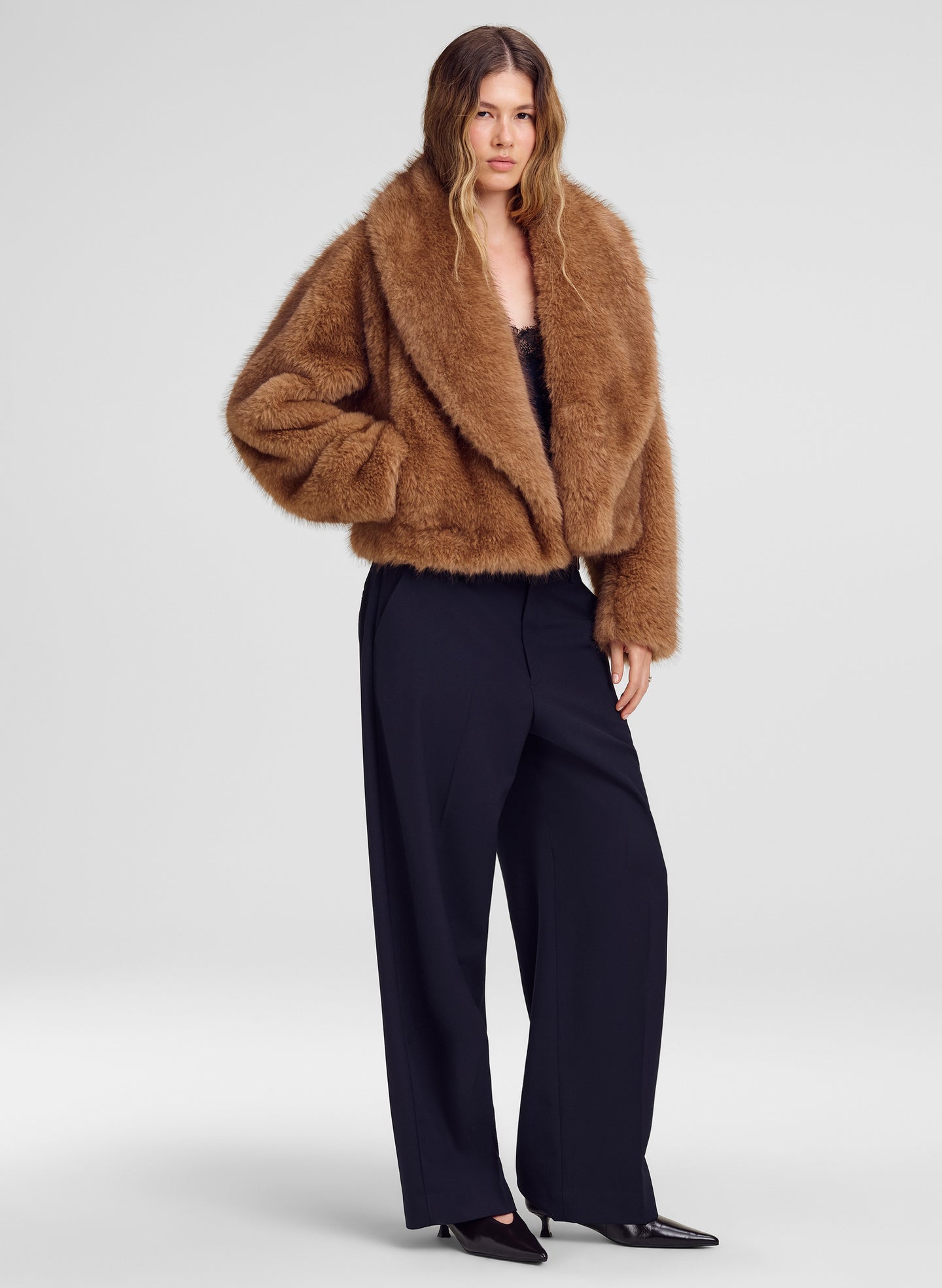 A model wearing a cropped, plush brown faux fur jacket over a black top and navy wide-leg trousers, standing against a light gray studio background.