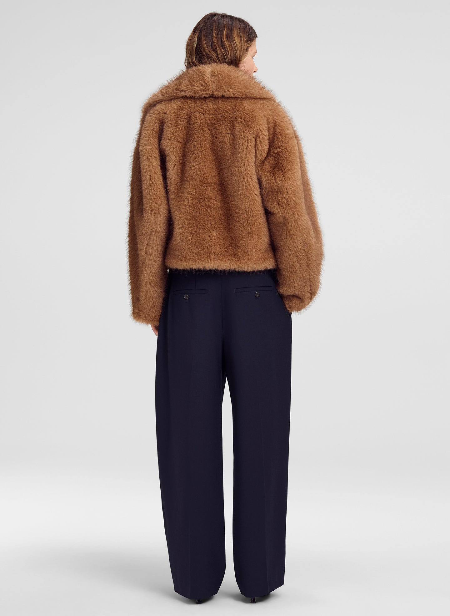 A model shown from the back wearing a cropped brown fur jacket and dark navy wide-leg trousers, photographed against a light gray studio background.