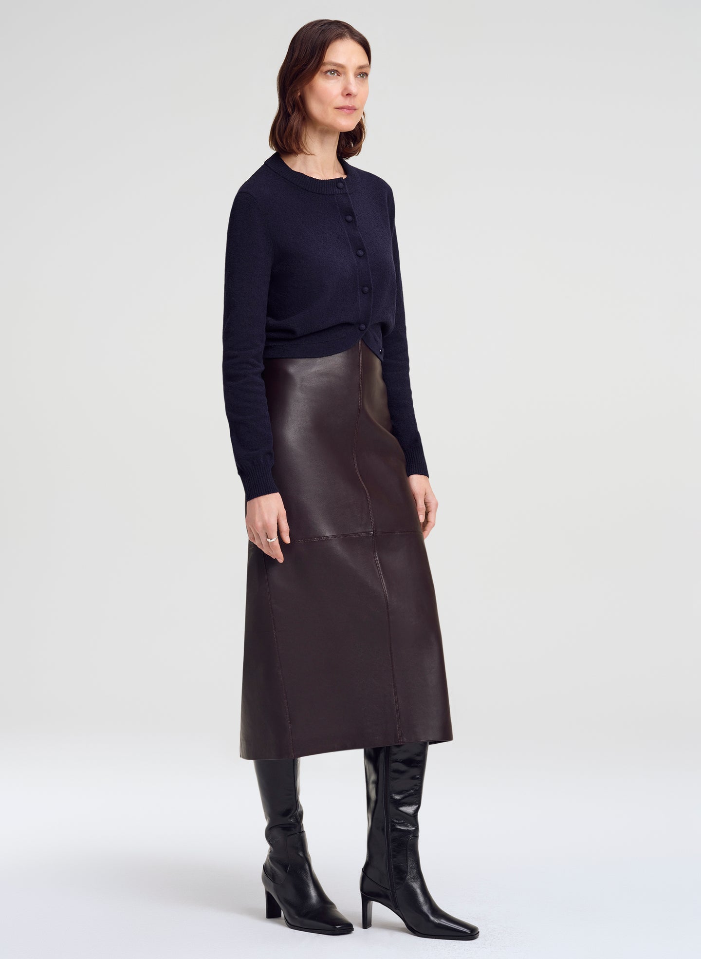 A woman modeling a dark navy long-sleeve button-front cardigan, a dark brown leather midi skirt, and black knee-high heeled boots against a light gray studio background.