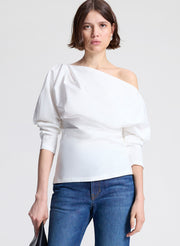 Woman wearing a white one-shoulder top and blue jeans on a light gray background