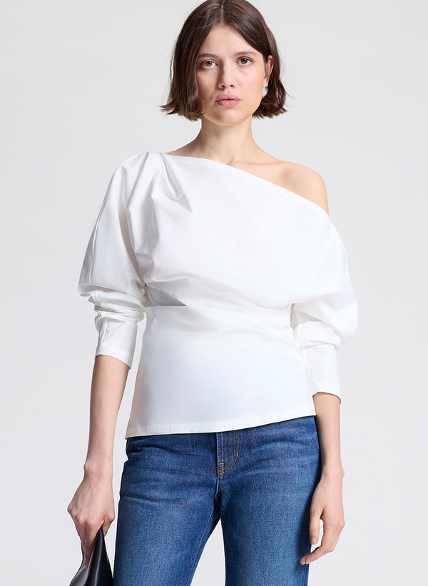 Woman wearing a white one-shoulder top and blue jeans on a light gray background