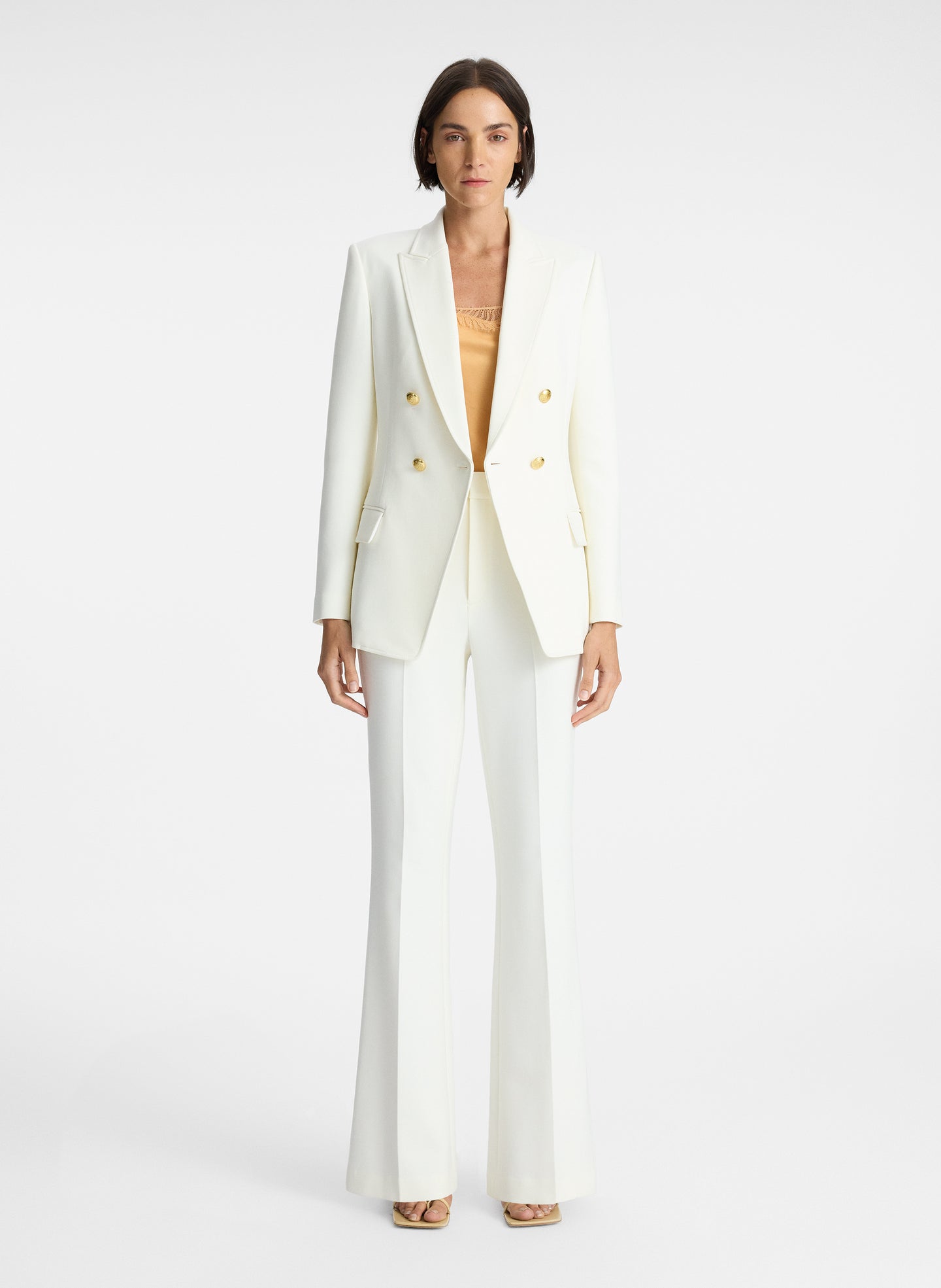 front view of woman wearing white blazer with gold buttons, beige camisole, and white pants