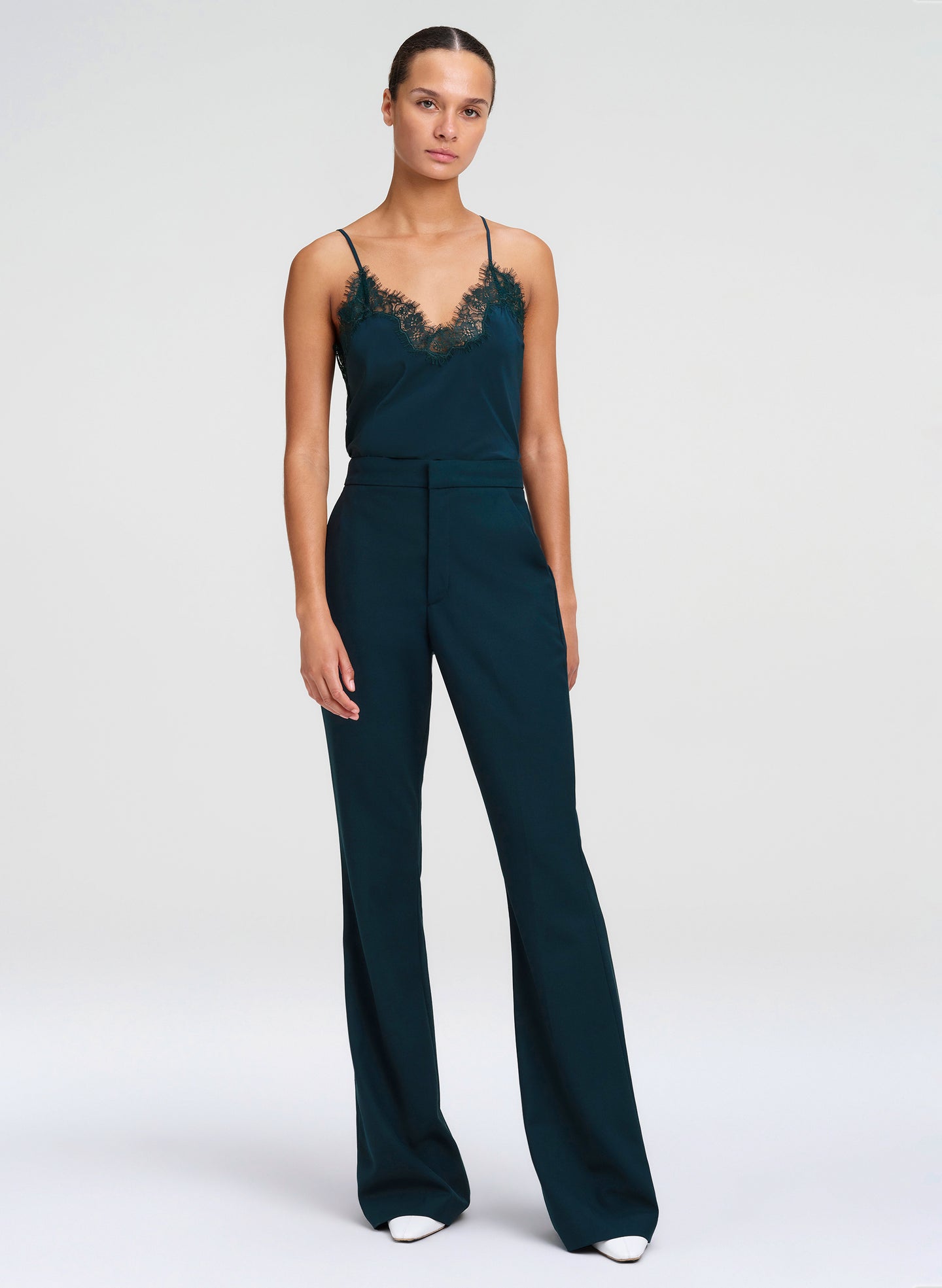 A model wearing a dark teal green outfit with a lace-trimmed camisole bodice and wide-leg trousers, standing against a light gray studio background.