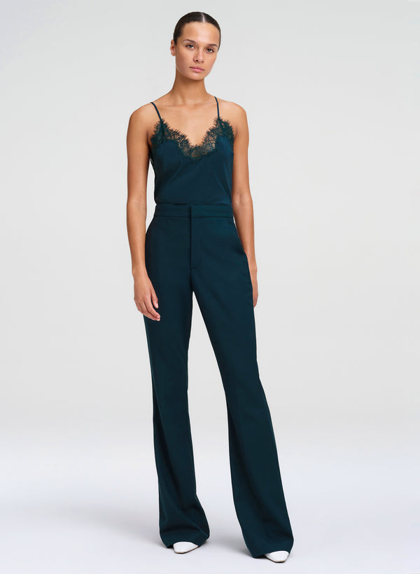 A model wearing a dark teal green outfit with a lace-trimmed camisole bodice and wide-leg trousers, standing against a light gray studio background.