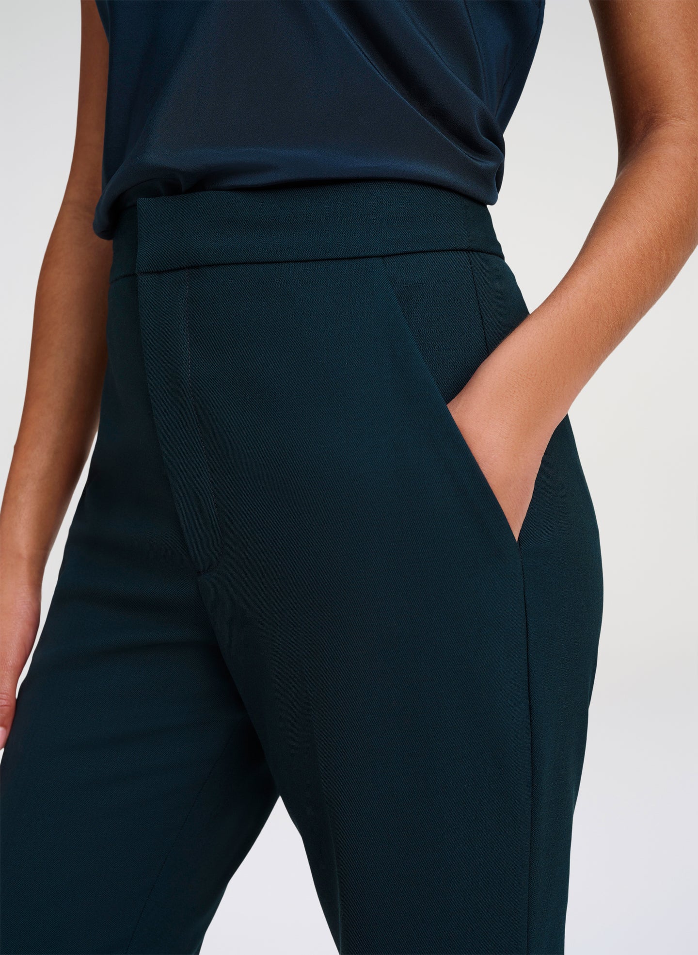 Close-up of a model wearing dark green high-waisted trousers with side pockets and a matching dark green top against a light gray studio background.