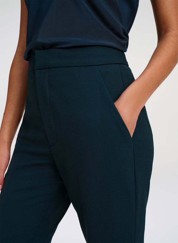 Close-up of a model wearing dark green high-waisted trousers with side pockets and a matching dark green top against a light gray studio background.