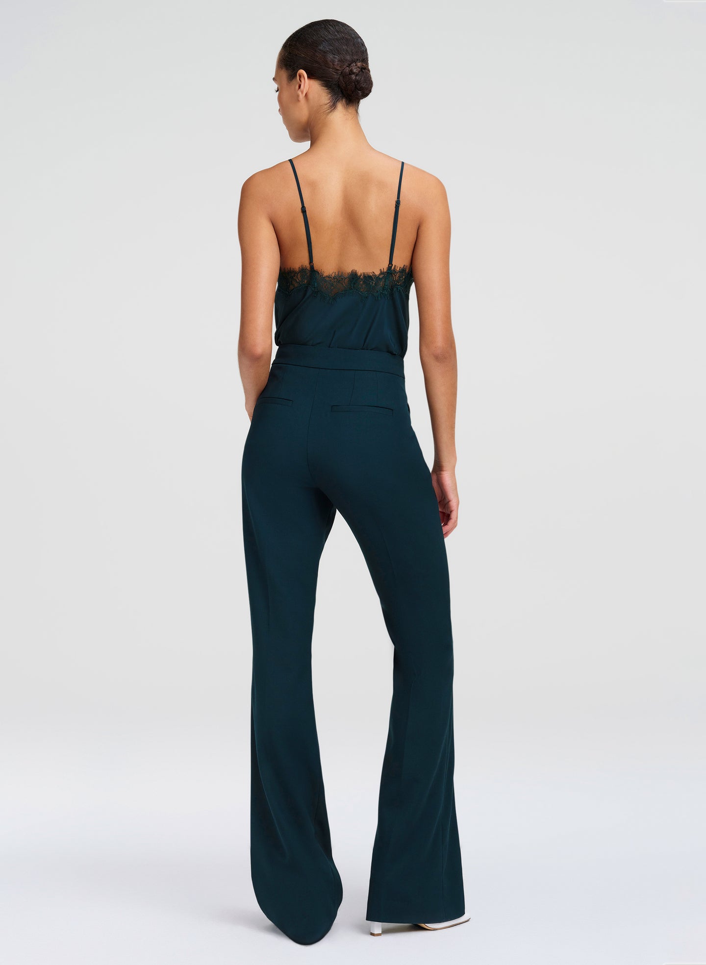 A woman shown from the back wearing a dark teal top with thin straps and a lace-trimmed back and pants, photographed against a light gray studio background.