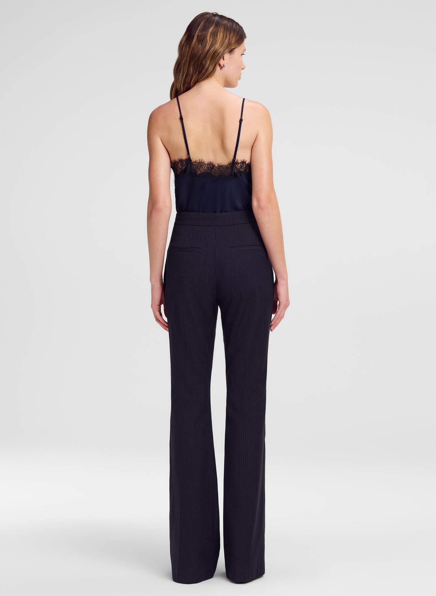 A woman viewed from the back wearing a dark navy sleeveless top with thin straps, a lace-trimmed neckline, and wide-leg pants, against a light gray studio background.