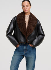 A model wearing a black faux-leather aviator-style jacket with a large brown shearling collar, paired with a beige top and blue jeans, against a light gray studio background.
