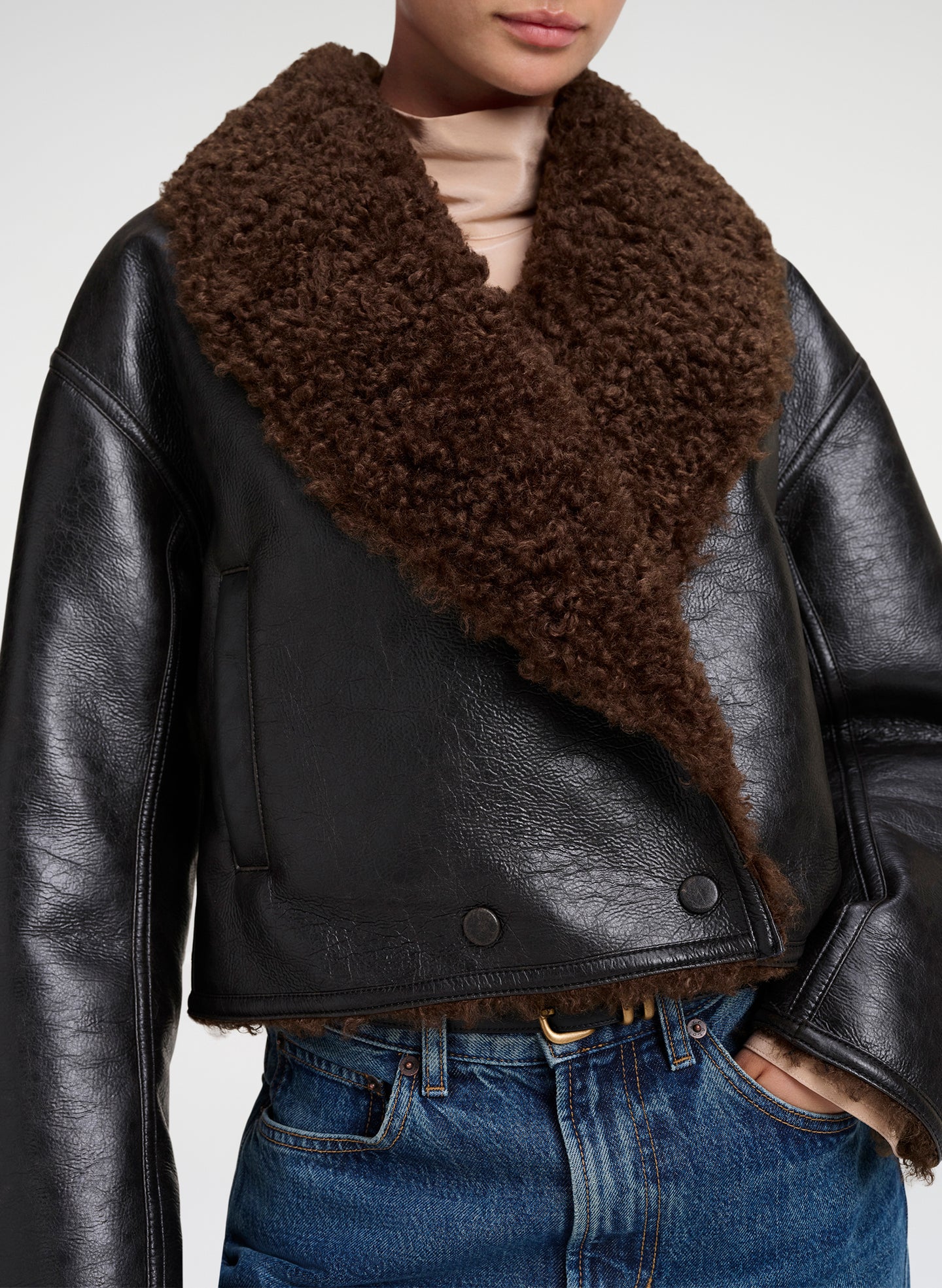 A cropped black leather jacket with a large, shaggy dark brown shearling-style collar, worn by a model with blue jeans against a light gray studio background.