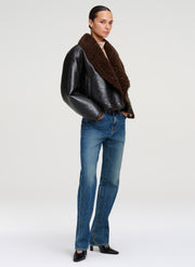 A model stands against a light gray studio background wearing a black faux leather jacket with a thick brown shearling collar, blue jeans, and black pointed shoes. 