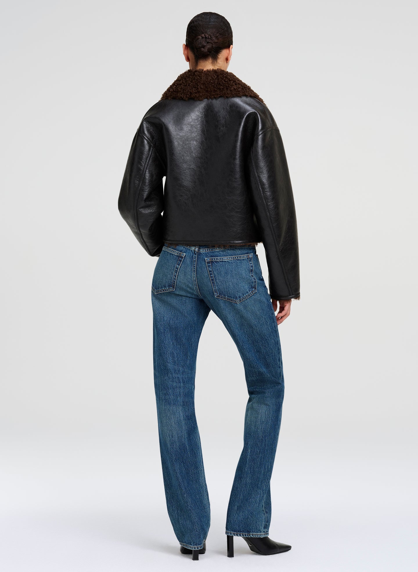 A model viewed from the back wearing a black faux-leather jacket with a brown shearling collar, paired with blue jeans and black heels, against a light gray studio background.