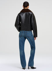 A model viewed from the back wearing a black faux-leather jacket with a brown shearling collar, paired with blue jeans and black heels, against a light gray studio background.