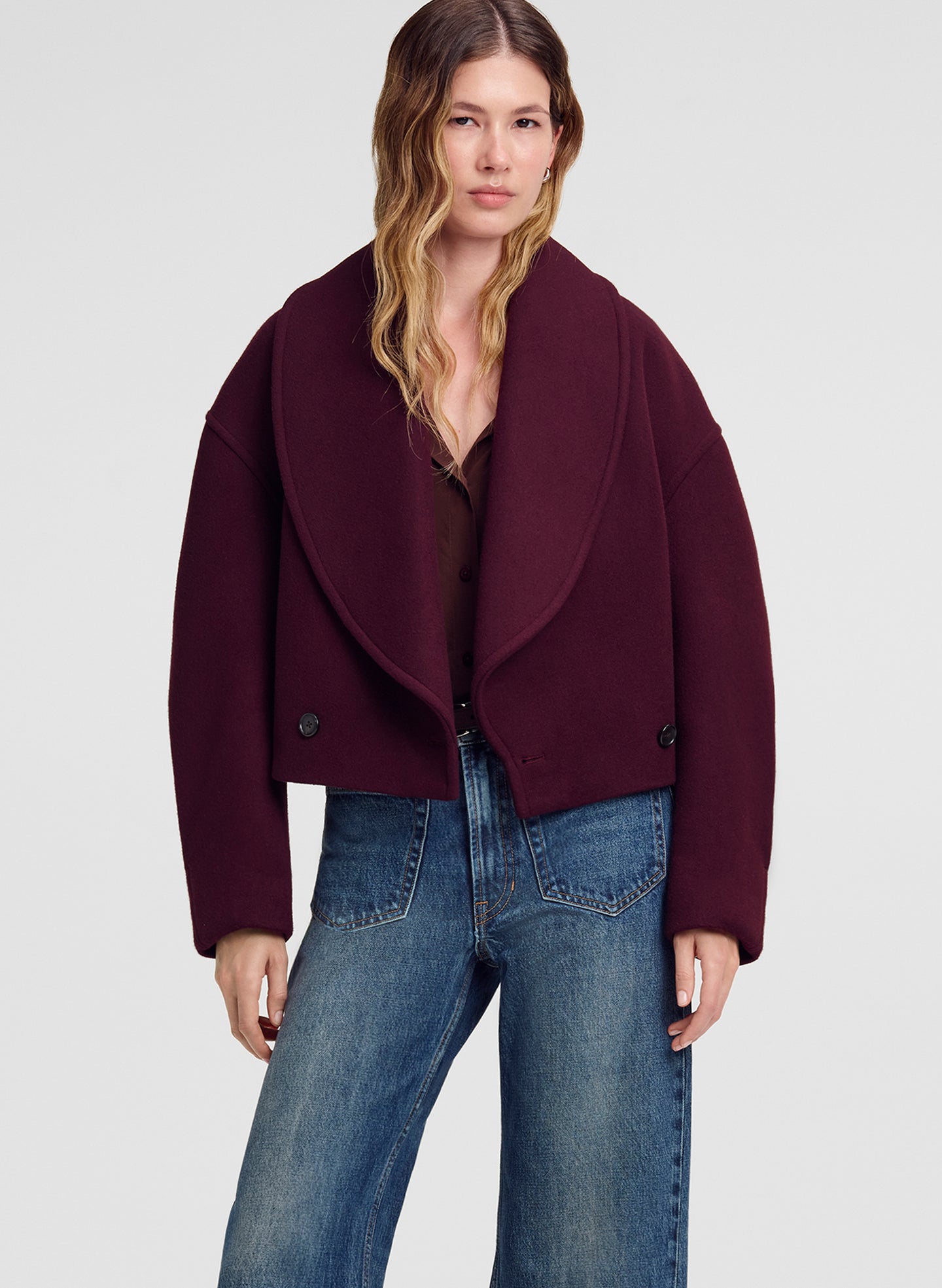 A woman modeling a cropped, oversized deep burgundy wrap jacket with dark buttons, worn over a dark top and blue jeans, against a light gray studio background.