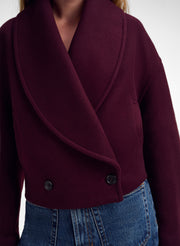 A close-up of a person wearing a deep burgundy wrap-style jacket with a wide shawl collar and two dark buttons, paired with blue jeans against a plain light background.