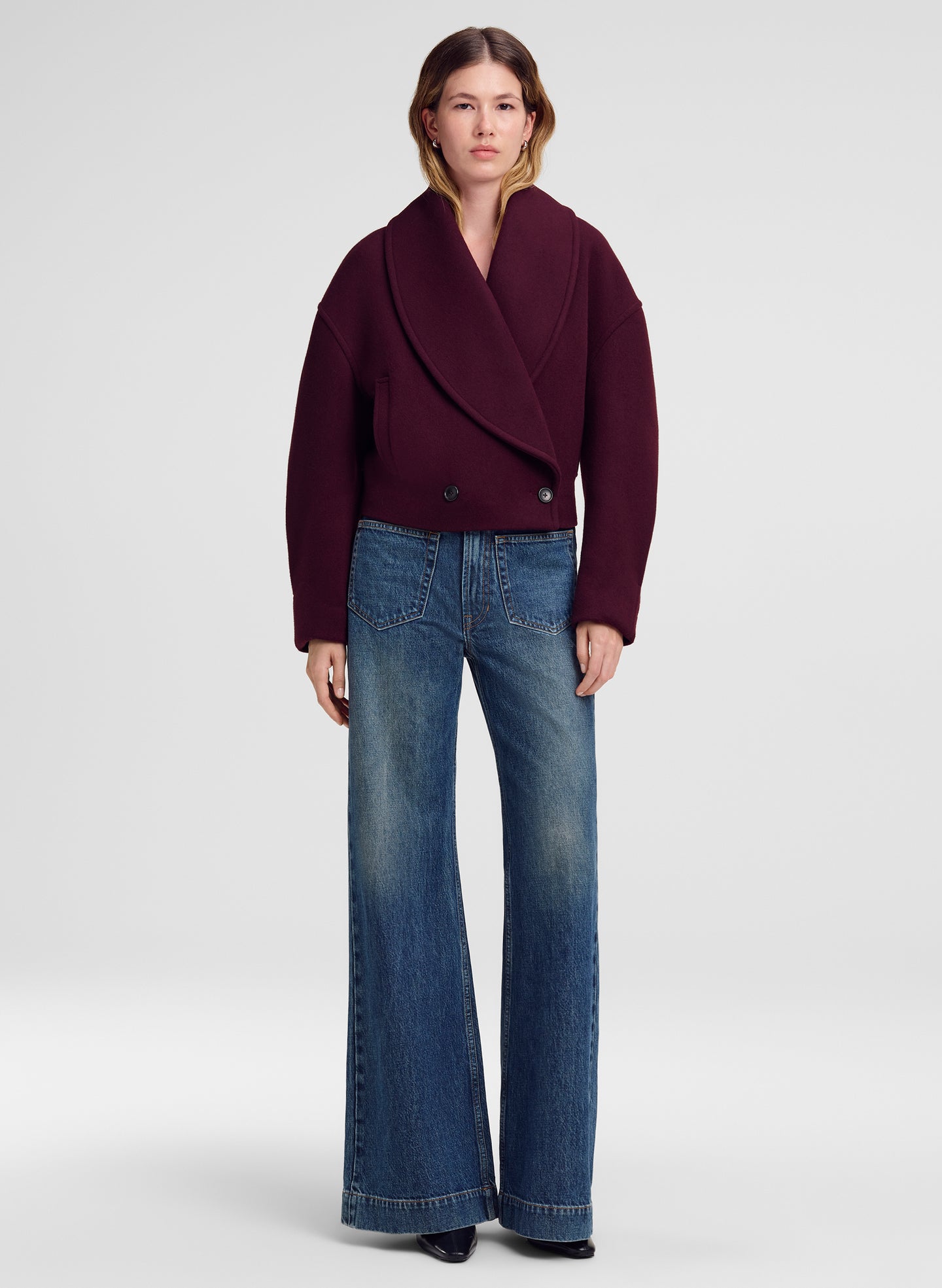 A model stands against a light gray studio background wearing a dark burgundy wrap-style jacket and blue wide-leg jeans.