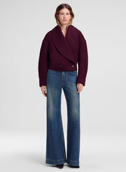 A model stands against a light gray studio background wearing a dark burgundy wrap-style jacket and blue wide-leg jeans.