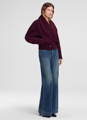 A woman modeling a deep burgundy cropped wrap jacket with wide lapels and blue flare jeans, standing against a light gray studio background.