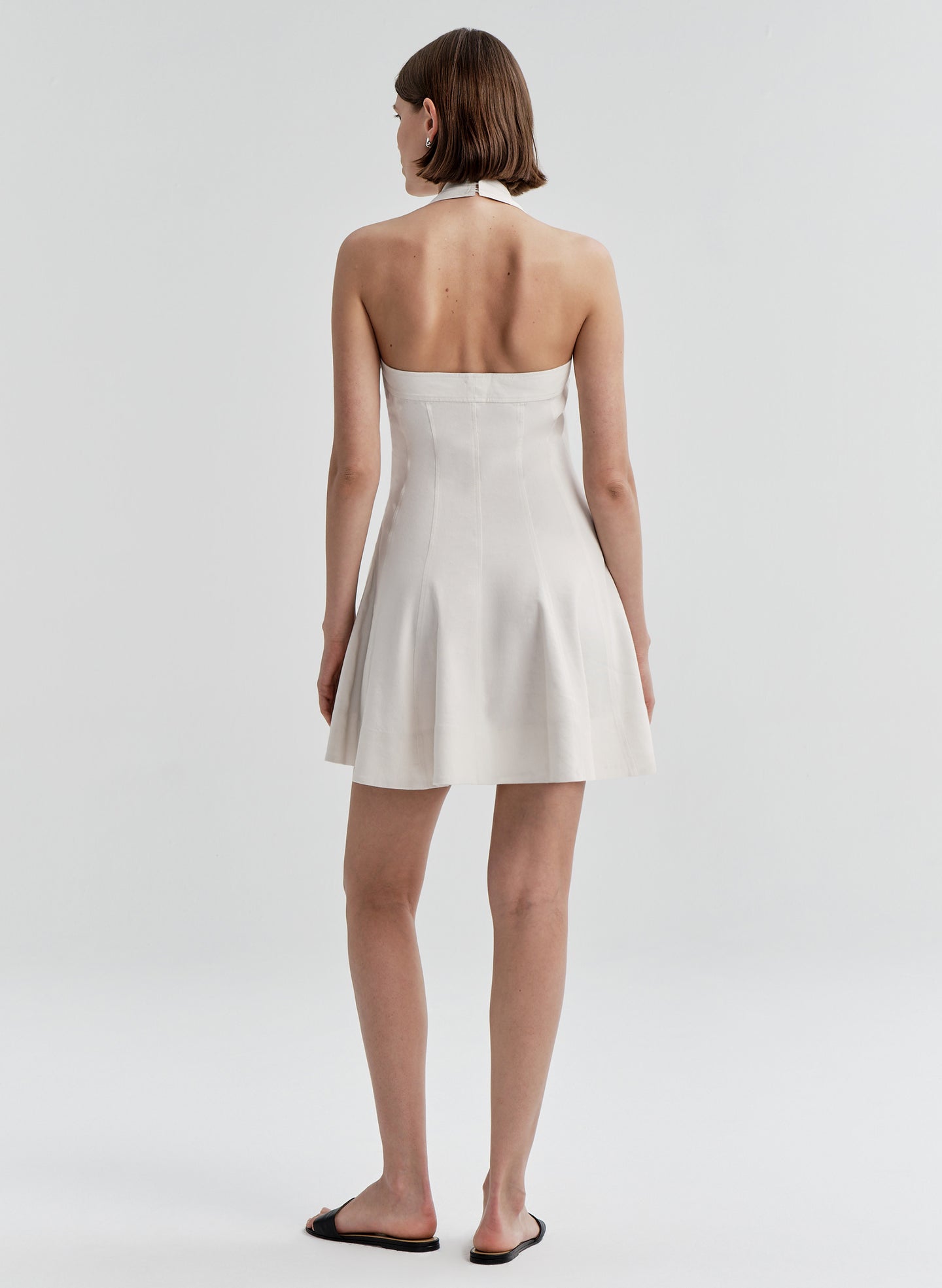 Woman wearing a beige linen halter neck dress with an open back on a plain background