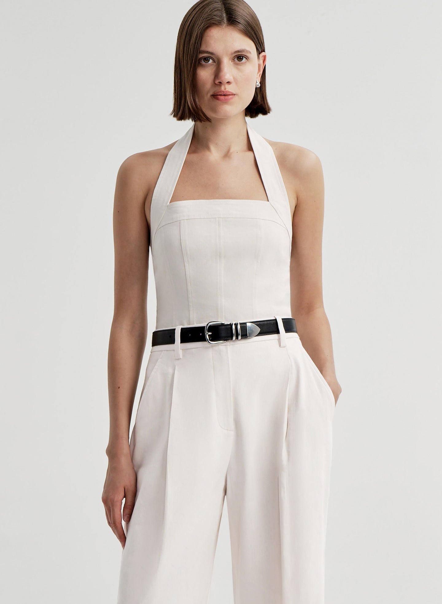 Woman wearing a cream colored halter top with a black belt with white pants and a light gray background