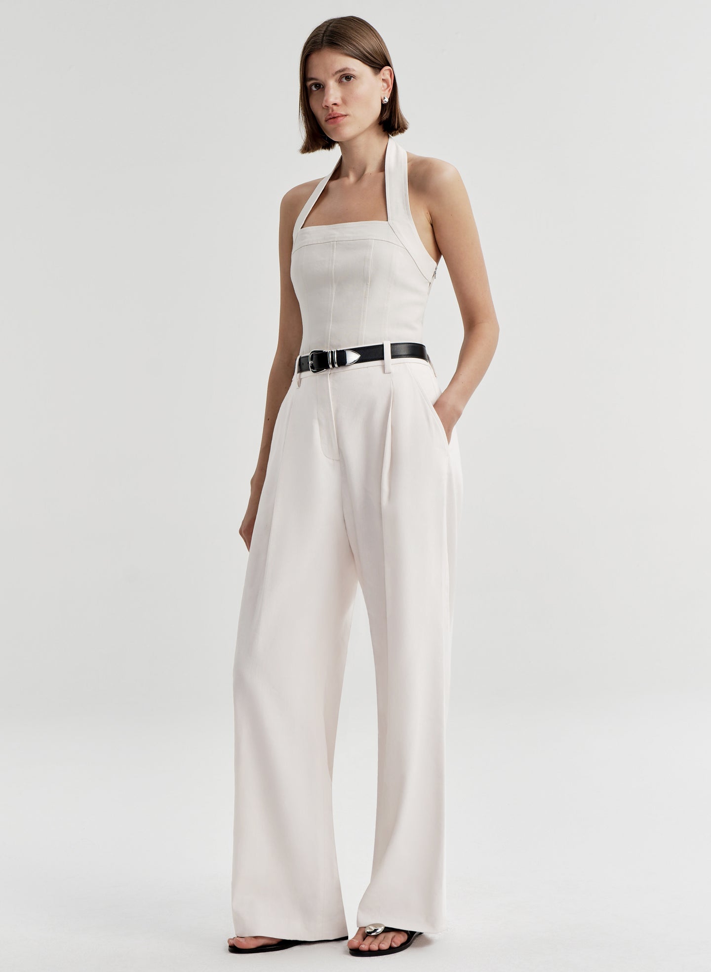 Woman wearing a cream colored halter top with a black belt with white pants and a light gray background