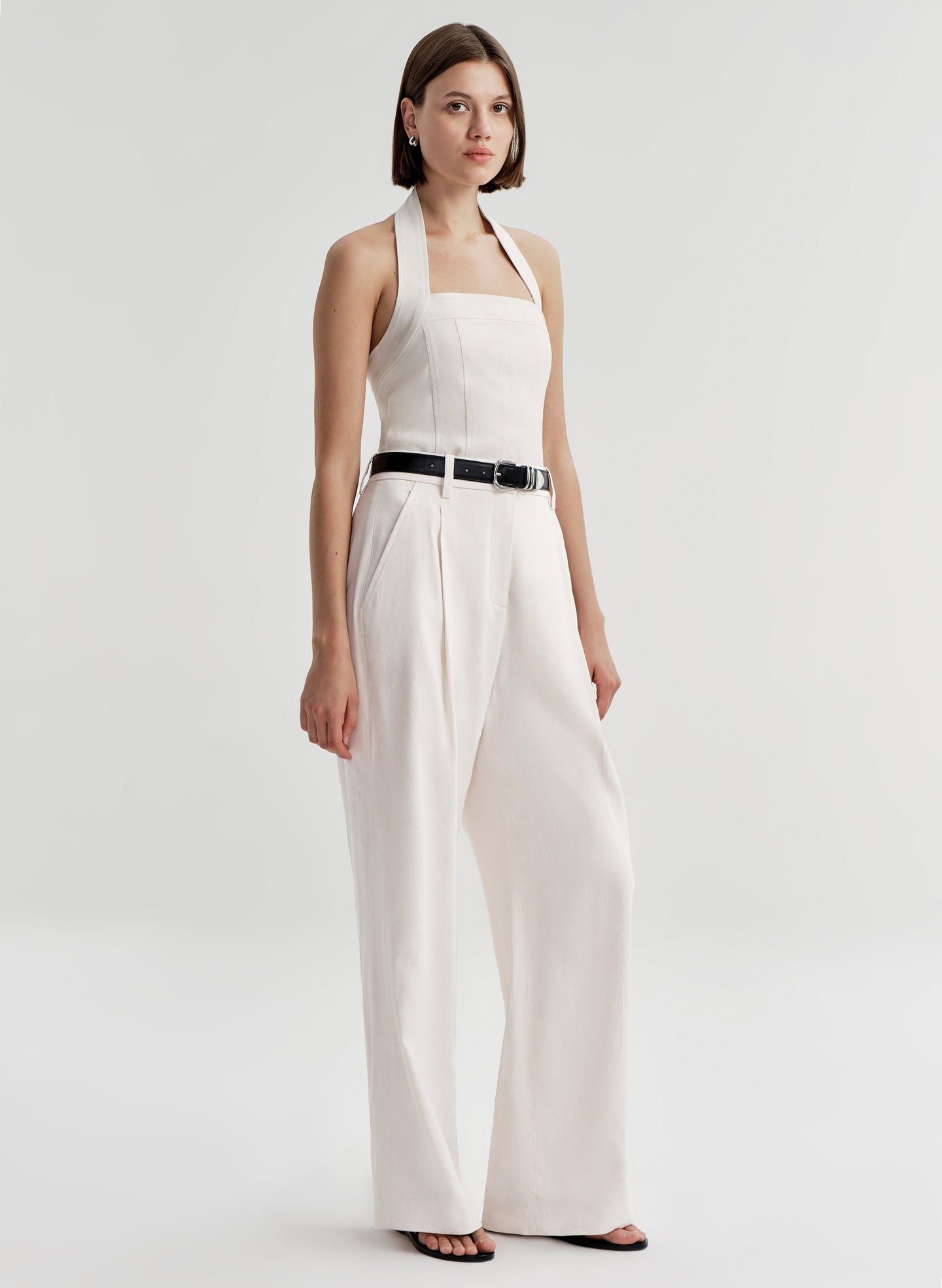 Woman wearing a cream colored halter top with a black belt with white pants and a light gray background