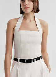 Woman wearing a cream colored halter top with a black belt with white pants and a light gray background