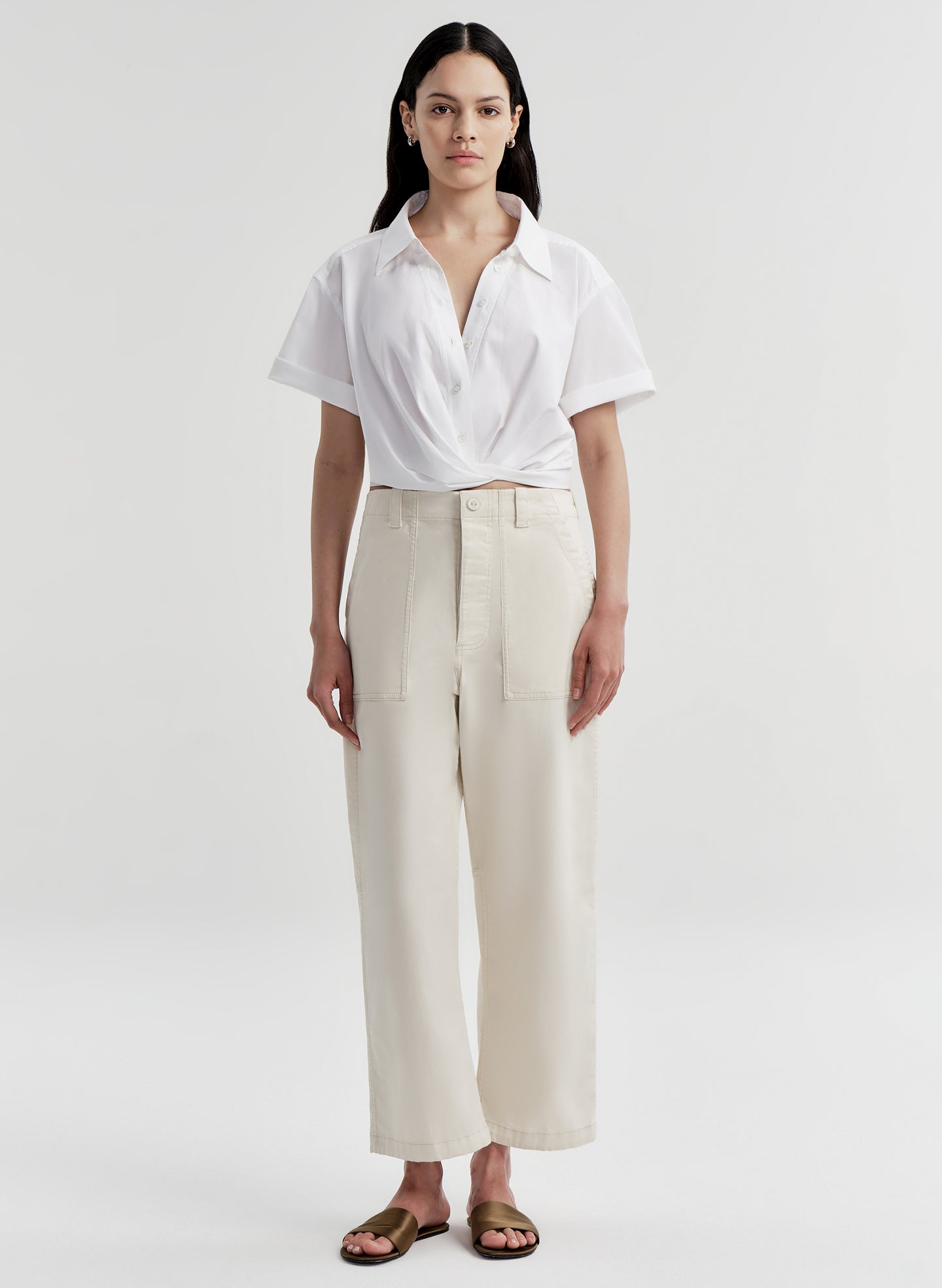 A woman stands against a light gray studio background wearing a white short-sleeve collared shirt with high-waisted cream wide-leg trousers and brown sandals.