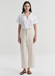 A woman stands against a light gray studio background wearing a white short-sleeve collared shirt with high-waisted cream wide-leg trousers and brown sandals.